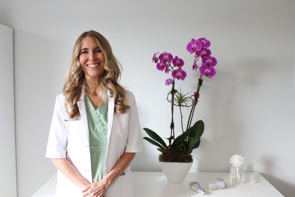 A smiling woman in a white lab coat stands beside a vase of orchids, radiating warmth and professionalism.