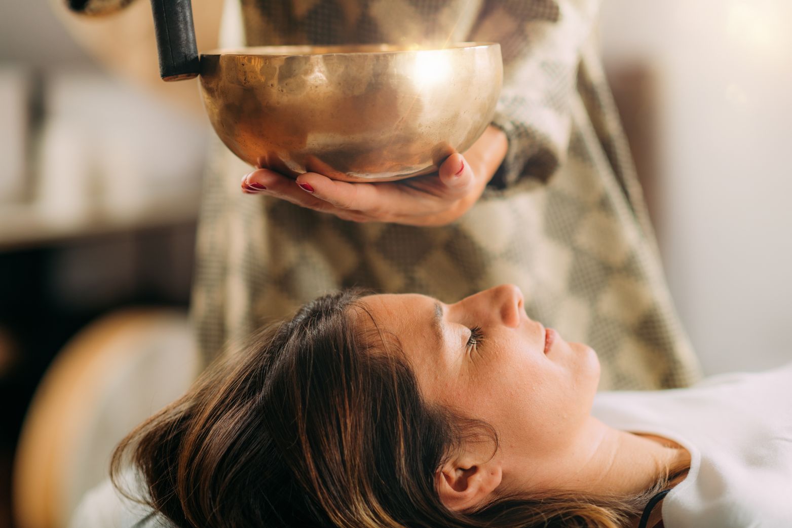 A woman lying down with her eyes closed while another person holds a gold Tibetan singing bowl above her.