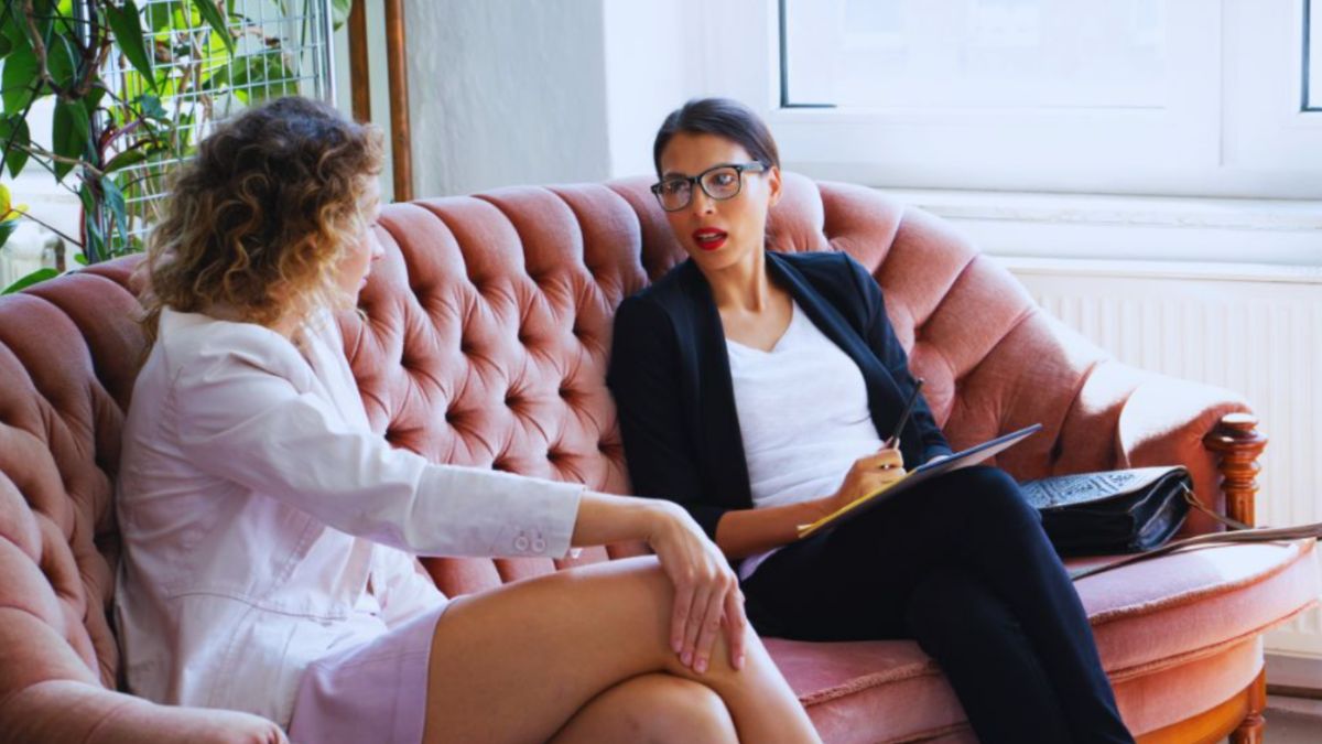 Career transition coaching session between two women sitting on a pink vintage couch. One woman, holding a clipboard, listens attentively while the other speaks, creating a professional and supportive environment.