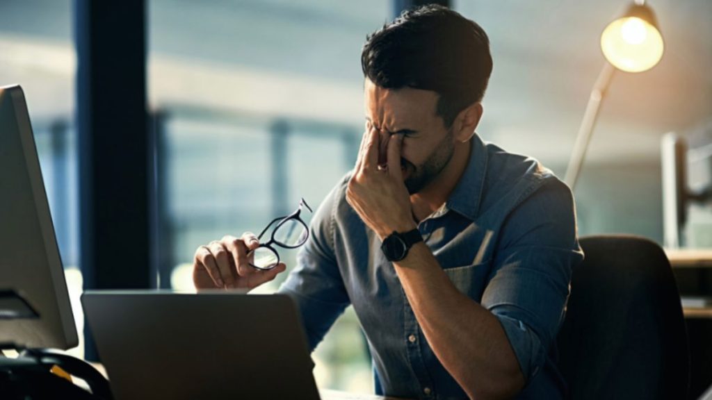 A stressed man at his desk, eyes closed with a hand on his face—capturing the emotional toll of burnout and the need for life coaching in Miami.
