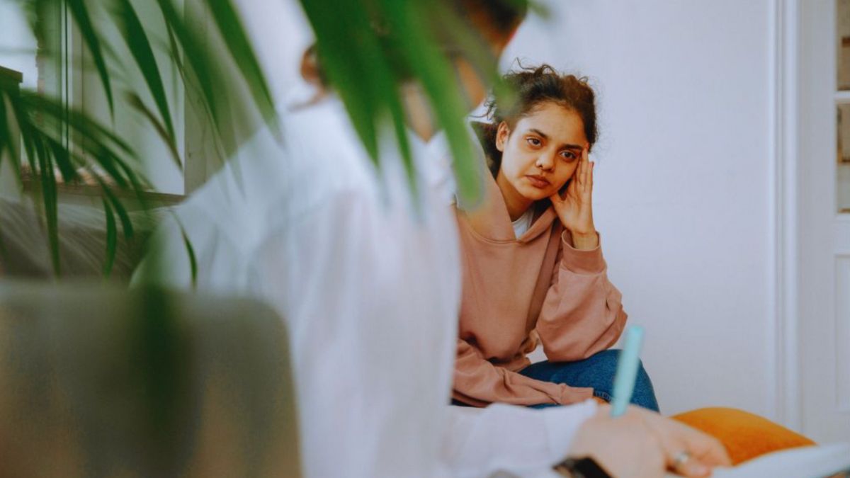 A young woman is sitting on a couch during a mindfulness coaching session, looking thoughtful and engaged, while the coach takes notes.