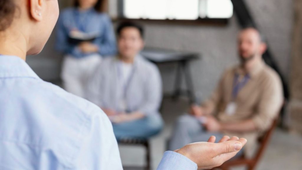 A person in a chair, hands extended, conversing with another seated individual about mindfulness coaching techniques.