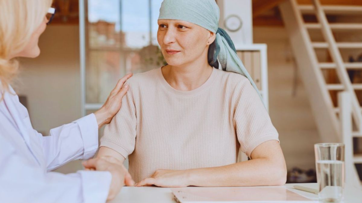 A woman in a soft-colored shirt and patterned headscarf sits at a table, engaged in a calm, supportive conversation with a professional. The professional gently places a hand on her shoulder, showing a calm conversation between two people, often representing life coaching after cancer recovery.