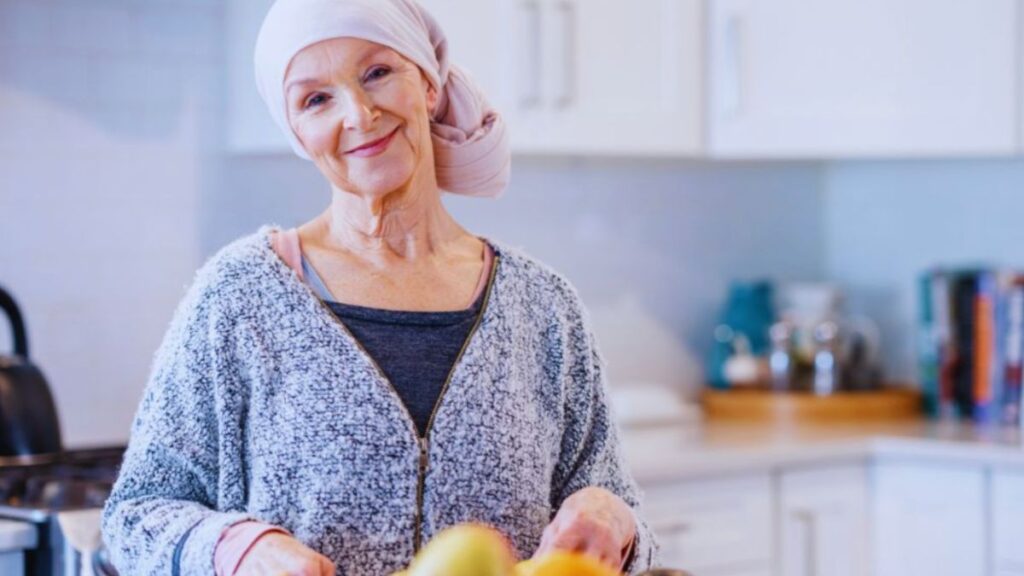 An older woman in a light pink headscarf and gray zip-up sweater prepares fruit at a kitchen counter. The bright, modern setting reflects a peaceful and supportive environment, capturing a calm moment in her recovery journey.