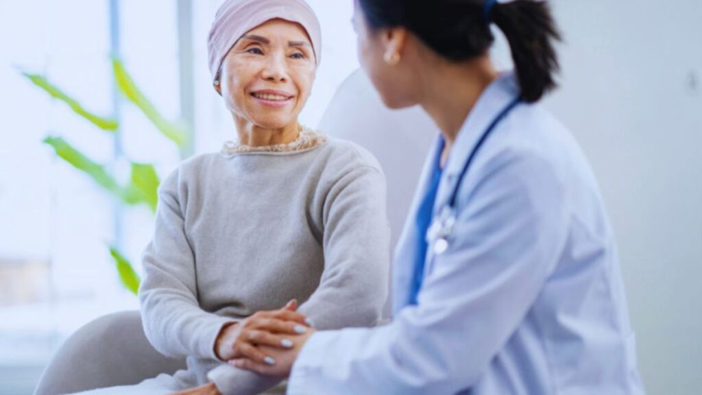 A healthcare provider and a cancer survivor engage in a supportive conversation in a bright, modern clinical setting. The provider, wearing a white coat and stethoscope, gently holds the survivor’s hand, reflecting compassion, encouragement, and non-clinical life coaching during post-treatment recovery.