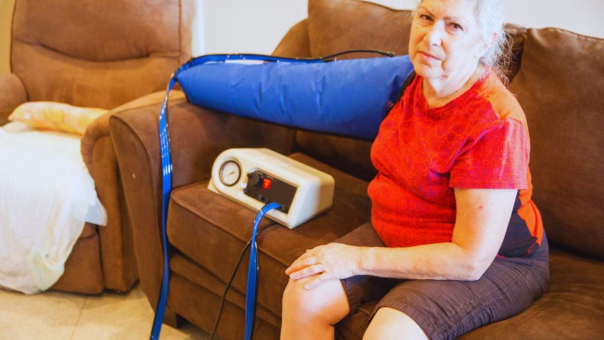 An older woman sits on a couch wearing a pneumatic compression sleeve on her arm connected to a home pneumatic compression device (PCD), receiving therapy in a home setting.